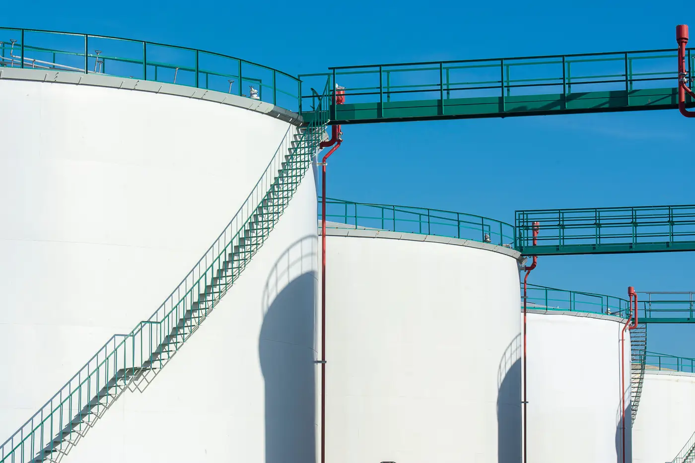 Row of large steel storage tanks with elevated walkways under a clear blue sky.