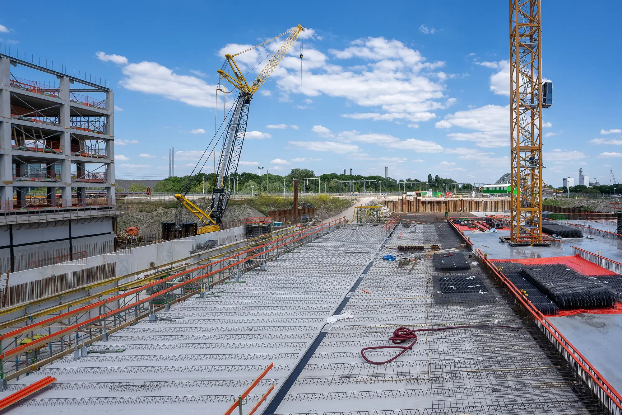 A reinforced concrete deck being laid on a large construction site, with tower and crawler cranes behind it under a blue sky